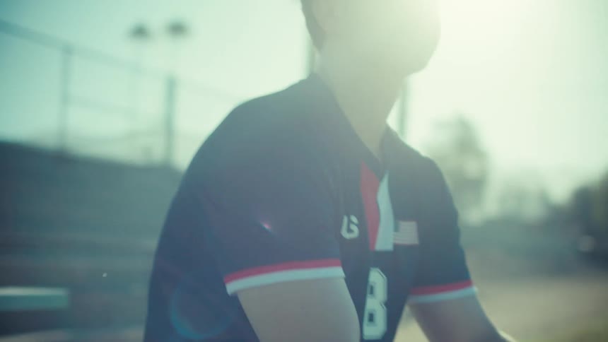 A pensive young soccer player in a USA jersey sits on a bench holding a soccer ball under the bright sun