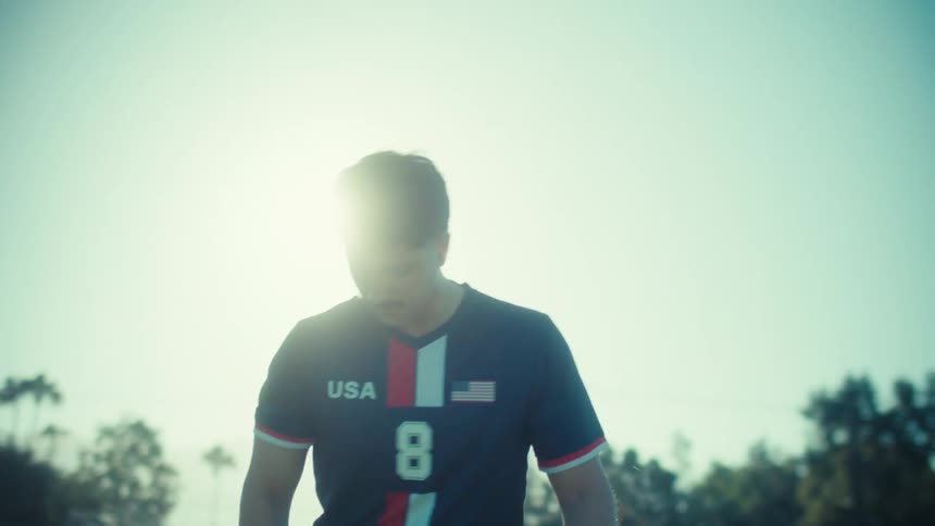A male soccer player in a USA jersey practices juggling the ball on a green field as the sun flares behind him