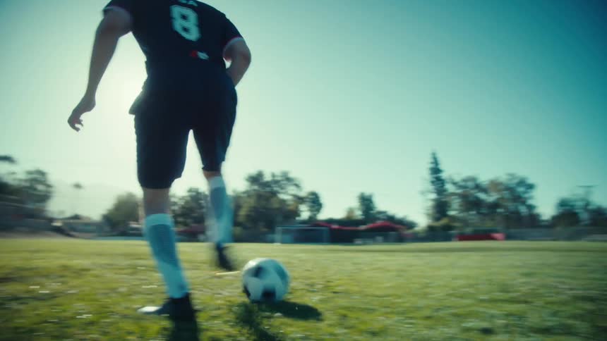 Slow-motion tracking shot of a male soccer player dribbling the ball down an empty field on a sunny day