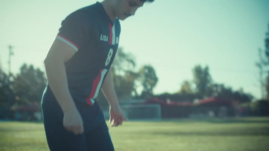 A young male soccer player in a USA uniform juggles a soccer ball on a sunny field in cinematic slow motion