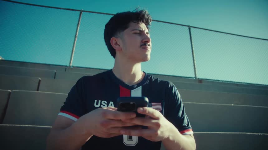 A young soccer player wearing a USA jersey sits on the bleachers and uses his phone before a game on a sunny day