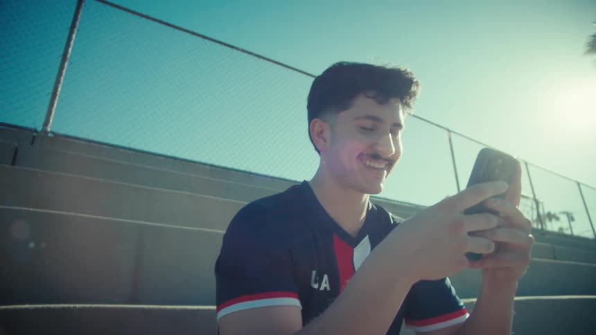 A young soccer player sits on the bleachers on a sunny day and smiles while using his smartphone