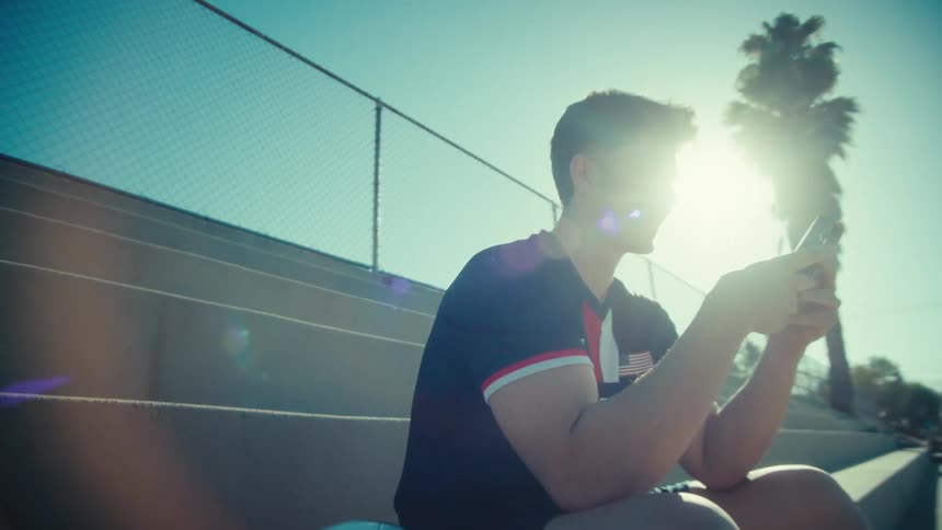 A smiling male soccer player uses his smartphone while sitting on stadium bleachers on a bright, sunny day