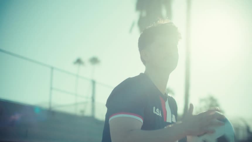 A focused young male soccer player in a USA jersey sits on the bleachers before standing up, looking out with determination as the sun flares brightly behind him