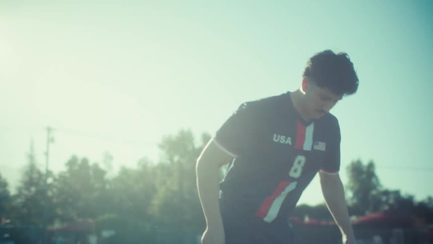 A skilled young soccer player in a USA jersey juggles a soccer ball in slow motion on a sunlit grass field
