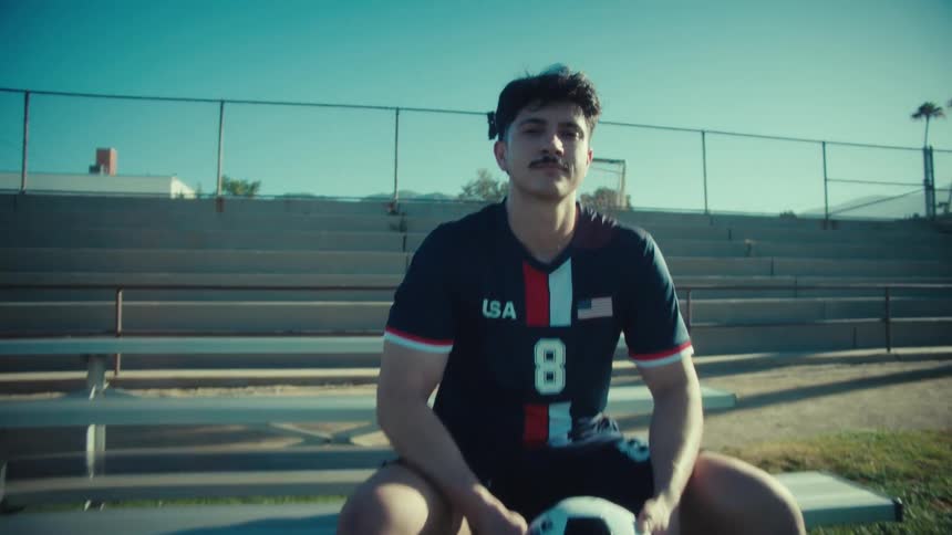 A young male soccer player in a USA uniform sits on a bench holding a ball, looking thoughtfully at the camera on a sunny day