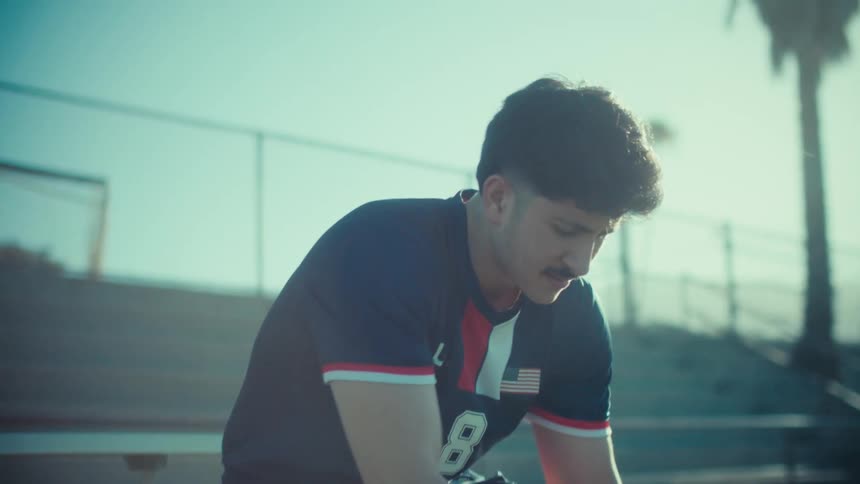 A young male soccer player in a USA uniform sits on a bench on a sunny field, preparing for the game