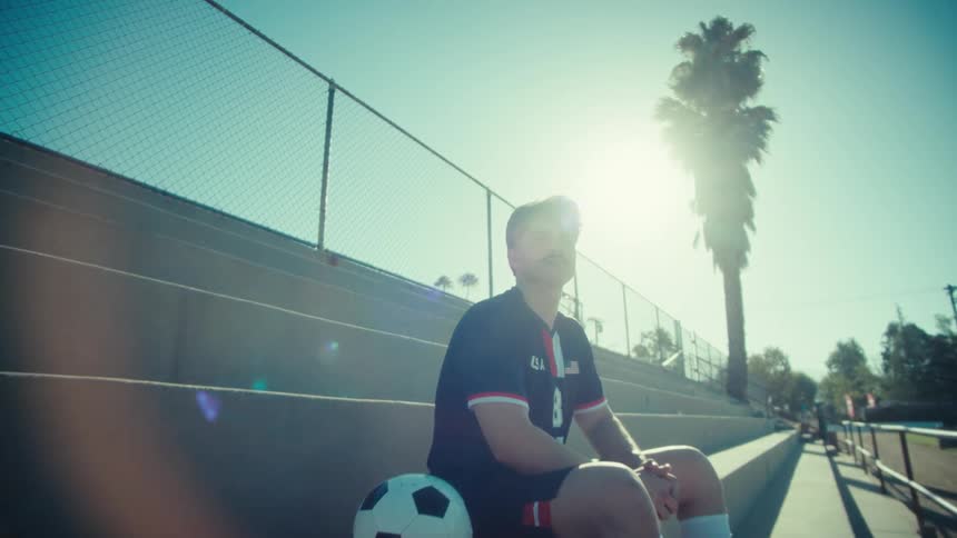 A slow push-in on a young, determined soccer player sitting on sunny bleachers and looking towards the sky with ambition