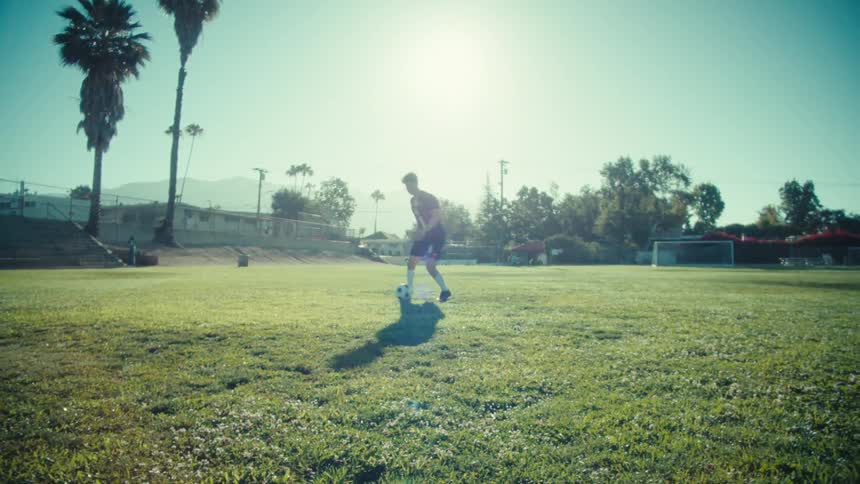 A soccer player skillfully dribbles a ball down a sunlit grass field in slow motion