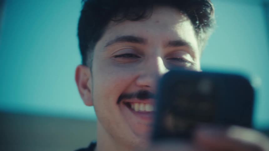 A low-angle close-up of a happy young man with a mustache smiling as he scrolls through his smartphone on a sunny day