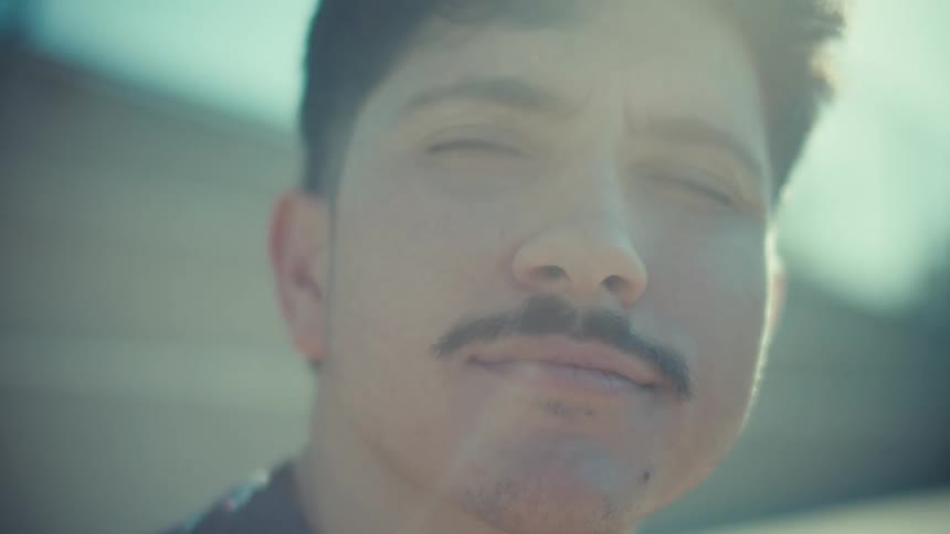 A dreamy close-up of a young man with a mustache basking in the sunlight and smiling peacefully
