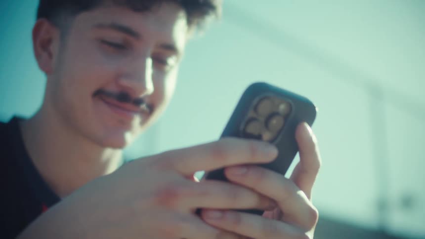 Close-up of a smiling young man with a mustache looking at his smartphone on a sunny day