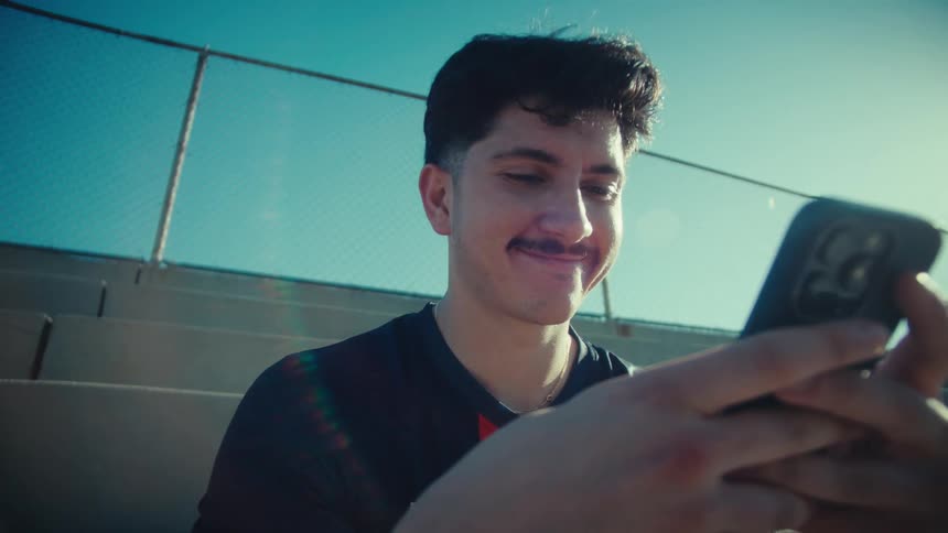 Low angle shot of a young man in a soccer jersey smiling as he scrolls on his smartphone while sitting on bleachers