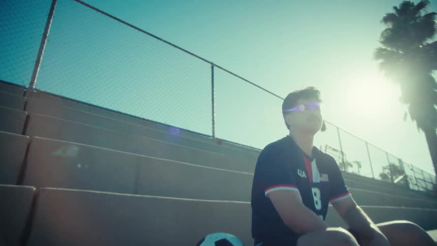 A determined young soccer player with a mustache sits alone on concrete bleachers on a sunny day, mentally preparing for the game