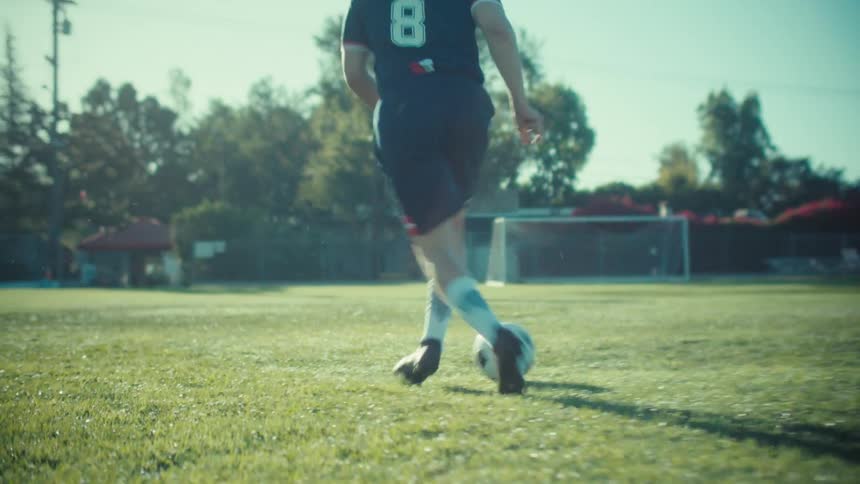 A close-up slow-motion shot follows from behind as a soccer player expertly dribbles a ball across a sun-drenched green field