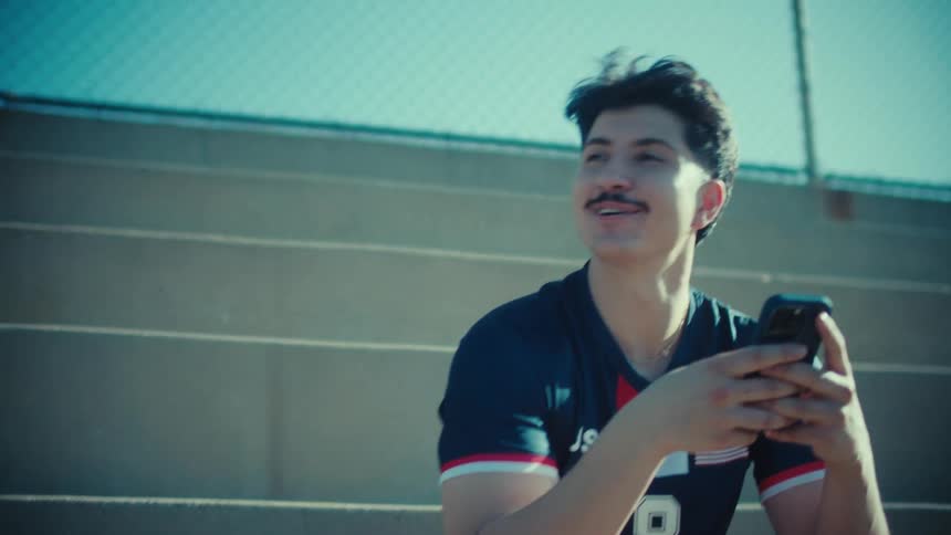 A happy young athlete sits on stadium bleachers, smiling and laughing while scrolling on his smartphone on a sunny day