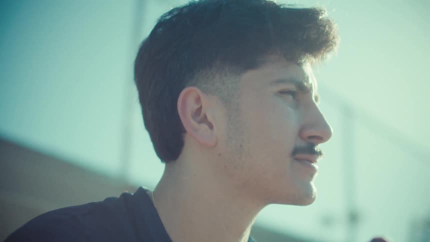A thoughtful young man in an athletic jersey scrolls through his smartphone while sitting outside in the bright sunlight