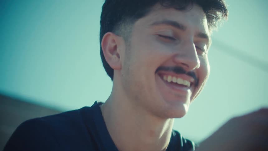A cheerful young soccer player with a mustache laughs while texting on his phone on sunny stadium bleachers
