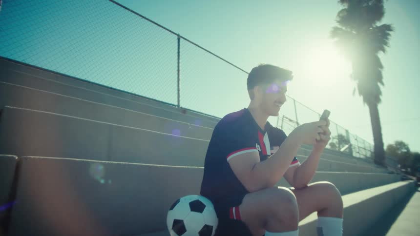 A smiling young soccer player takes a break on the bleachers, happily scrolling through his phone in the bright sunshine