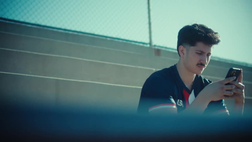 Slow zoom on a young male soccer player in a USA jersey sitting on sunny stadium bleachers and scrolling on his smartphone