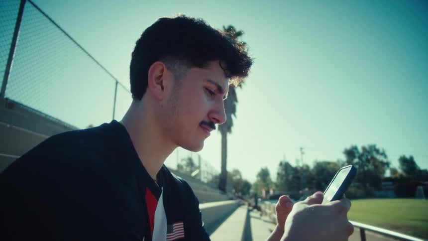 A cinematic close-up of a young male athlete sitting on the bleachers and scrolling through his phone on a sunny day