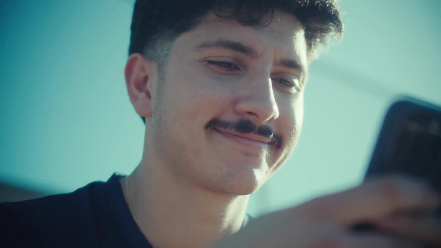 A low-angle close-up of a smiling young man with a mustache laughing while scrolling on his phone on a sunny day