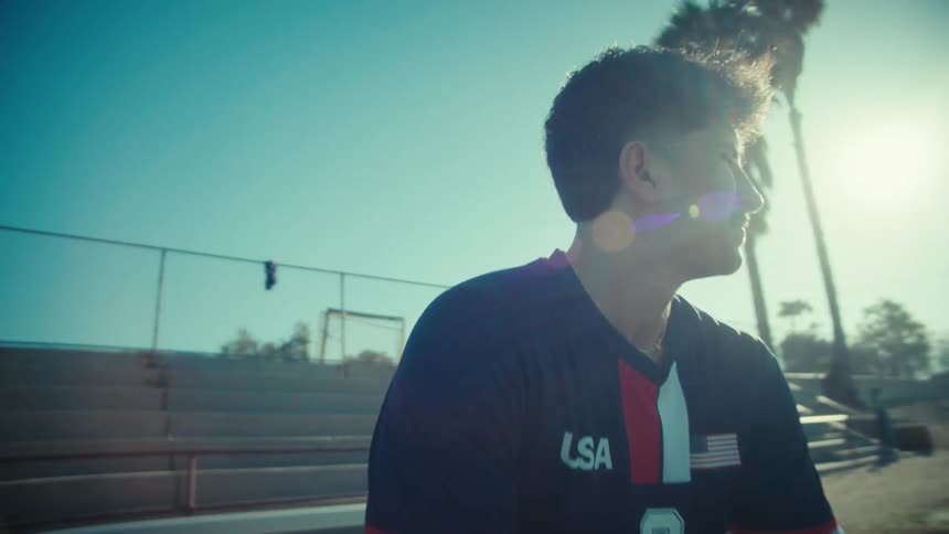 A determined young soccer player in a USA jersey sits contemplatively on the bleachers, looking out over the field on a sunny day