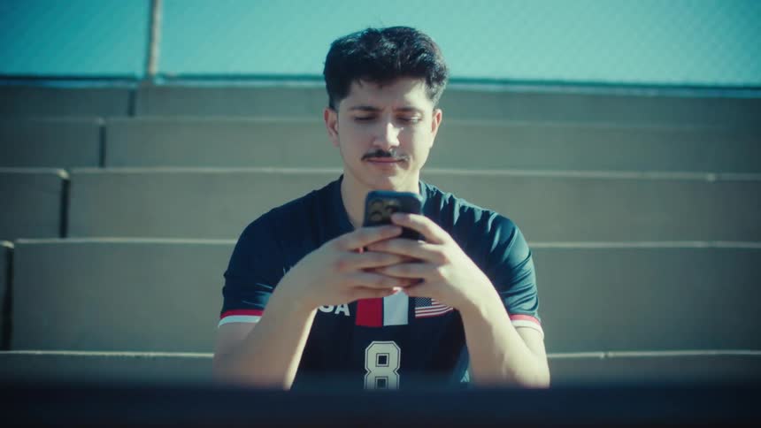A handsome young man in a USA soccer jersey smiles while scrolling on his smartphone as he sits on sunny stadium bleachers