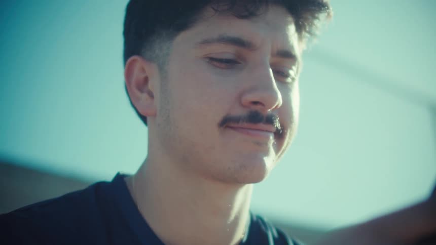 A young man with a mustache smiles and laughs while looking at his smartphone outdoors in the bright sun
