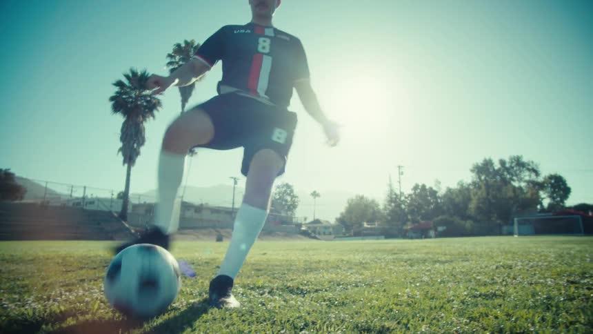 A male soccer player dribbles a ball in slow motion on a sunny day with palm trees in the background