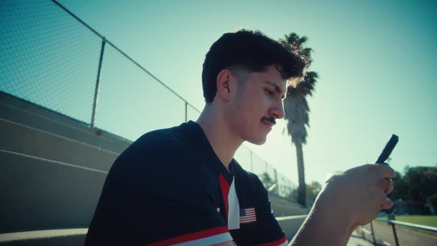 A young male athlete sits on bleachers on a sunny day, scrolling on his smartphone with a strong sun flare behind him