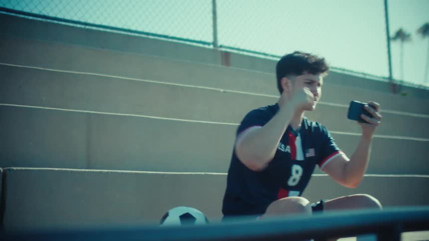 An ecstatic soccer player in full uniform celebrates a win on his mobile game while sitting on sunny stadium bleachers