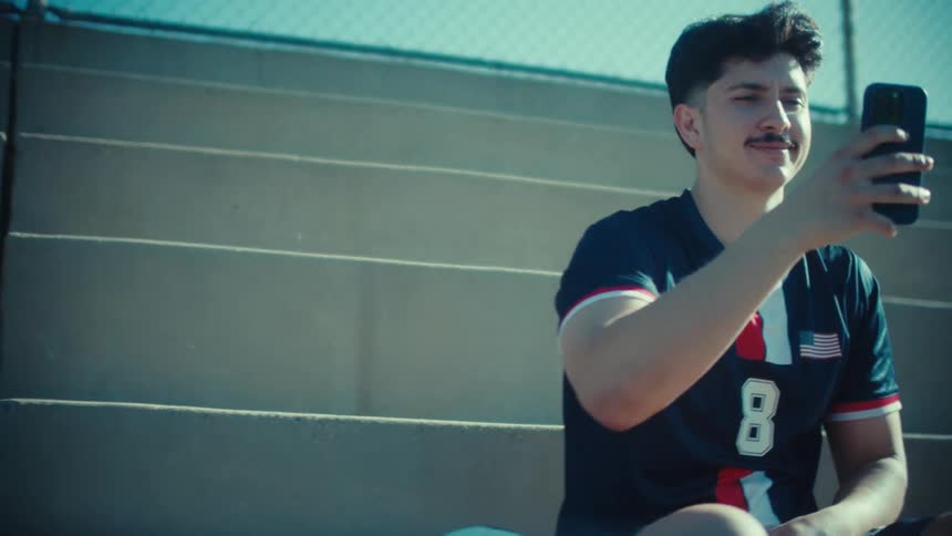 A young soccer player sits on the bleachers and happily video chats on his phone on a sunny day