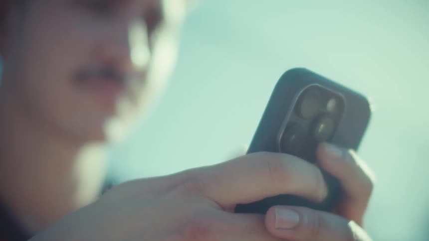 A cinematic close-up of a young man with a mustache using his smartphone outdoors on a bright, sunny day