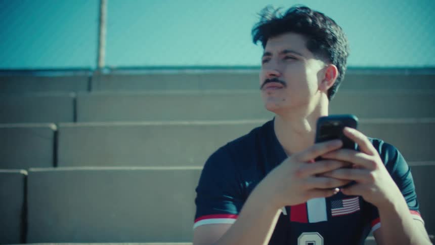 A young soccer player sits on the sunny bleachers of a stadium, intently focused on his smartphone