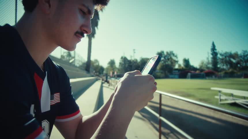 A young male athlete in a sports jersey sits in the bleachers on a sunny day scrolling through his smartphone