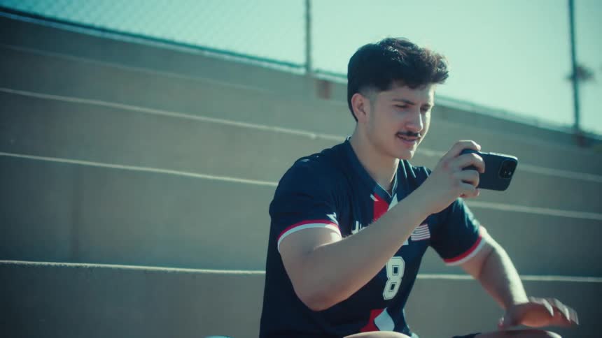 A young man in a USA soccer jersey excitedly plays a game on his phone, then stands up and celebrates a big win in the stadium bleachers