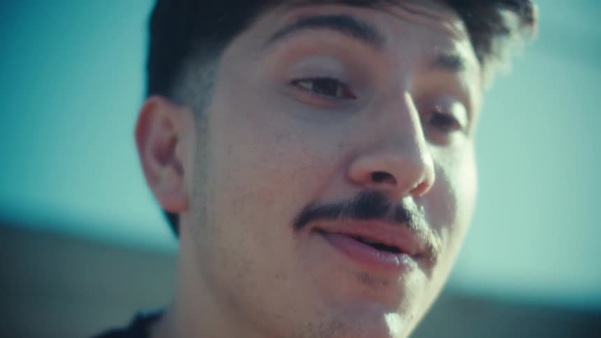 A close-up cinematic shot of a young man with a mustache smiling and talking on his smartphone in the bright sunlight