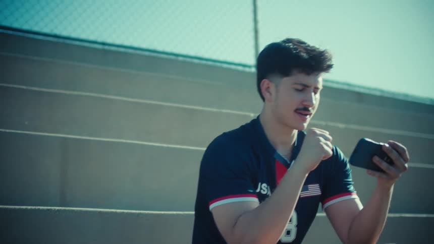 An excited young male soccer player in a USA jersey celebrates winning a mobile game while sitting on the stadium bleachers