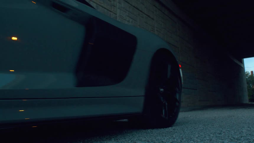 A low-angle view shows a sleek grey sports car with its headlights on driving out from under a dark, concrete overpass