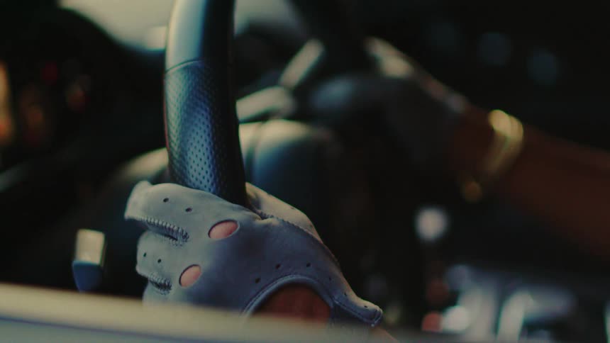 A close-up shot of a person's hand in a stylish grey driving glove gripping the steering wheel of a modern car