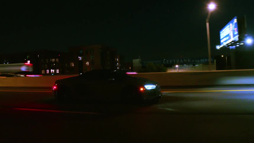 Side profile of a sleek grey sports car cruising down an urban highway at night with traffic in the background