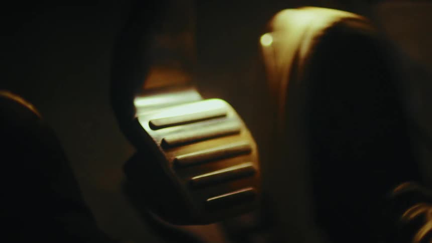 A gritty close-up of a driver's foot in a sneaker working the gas and brake pedals in a dimly lit car