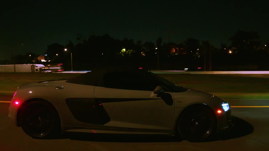 A sleek grey convertible sports car glides along a highway at night, its body illuminated by passing streetlights
