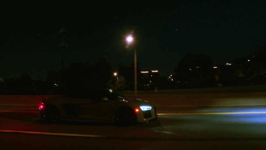 A sleek grey Audi R8 convertible cruises down an urban highway at night with its headlights illuminating the road