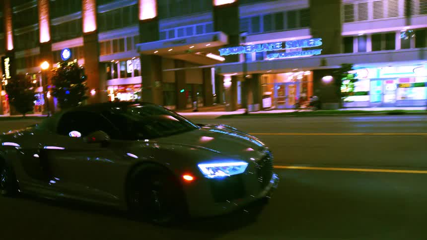 A luxury sports car cruises down an urban street at night, its headlights illuminating the road as it passes by city buildings