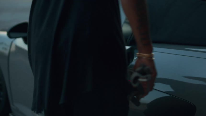 A man in black walks in slow motion towards his gray convertible sports car, gets in, and shuts the door