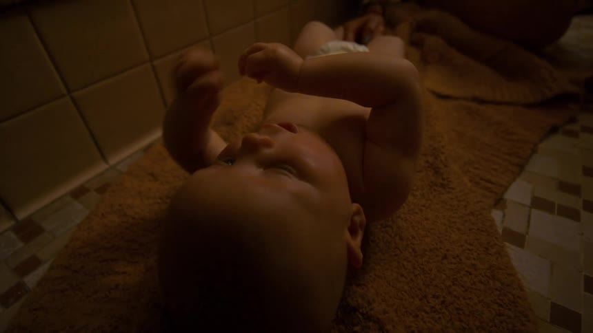 An adorable baby in a diaper lies on a towel on the bathroom floor, moving gently under a warm light