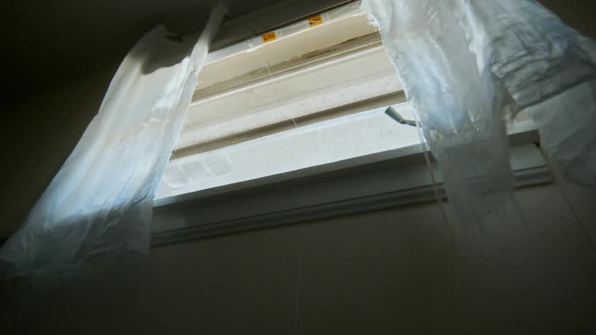 Torn white curtains blow eerily in the breeze next to a dusty window in a dark, abandoned room