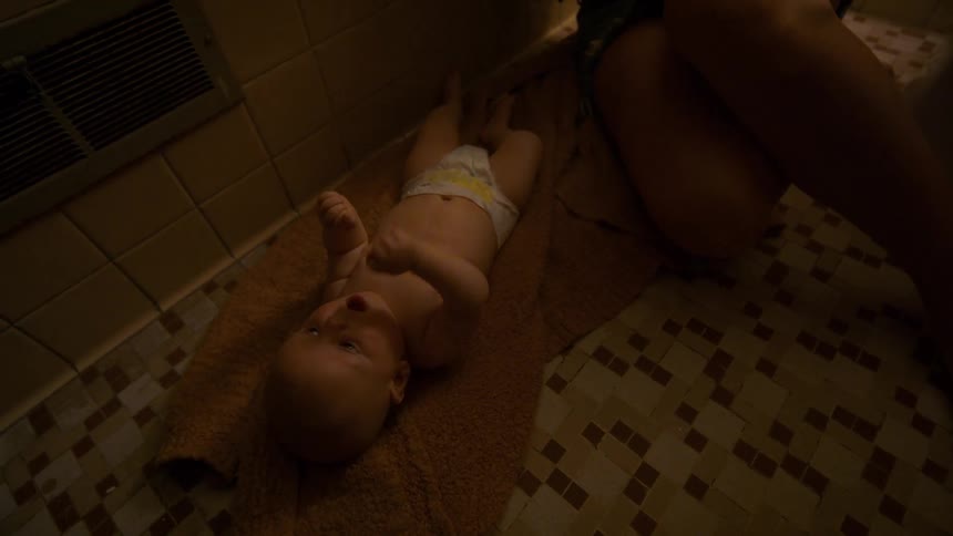 Baby lying on a towel in a dim bathroom while an adult kneels nearby during a diaper change